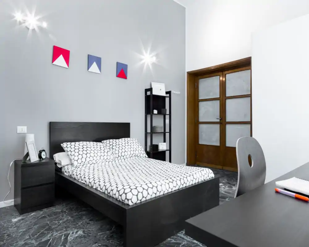 Dormitorio moderno y luminoso con cama de madera oscura y ropa de cama estampada, mesita y estantería; arte geométrico en la pared y zona de escritorio crean un espacio funcional y con estilo.