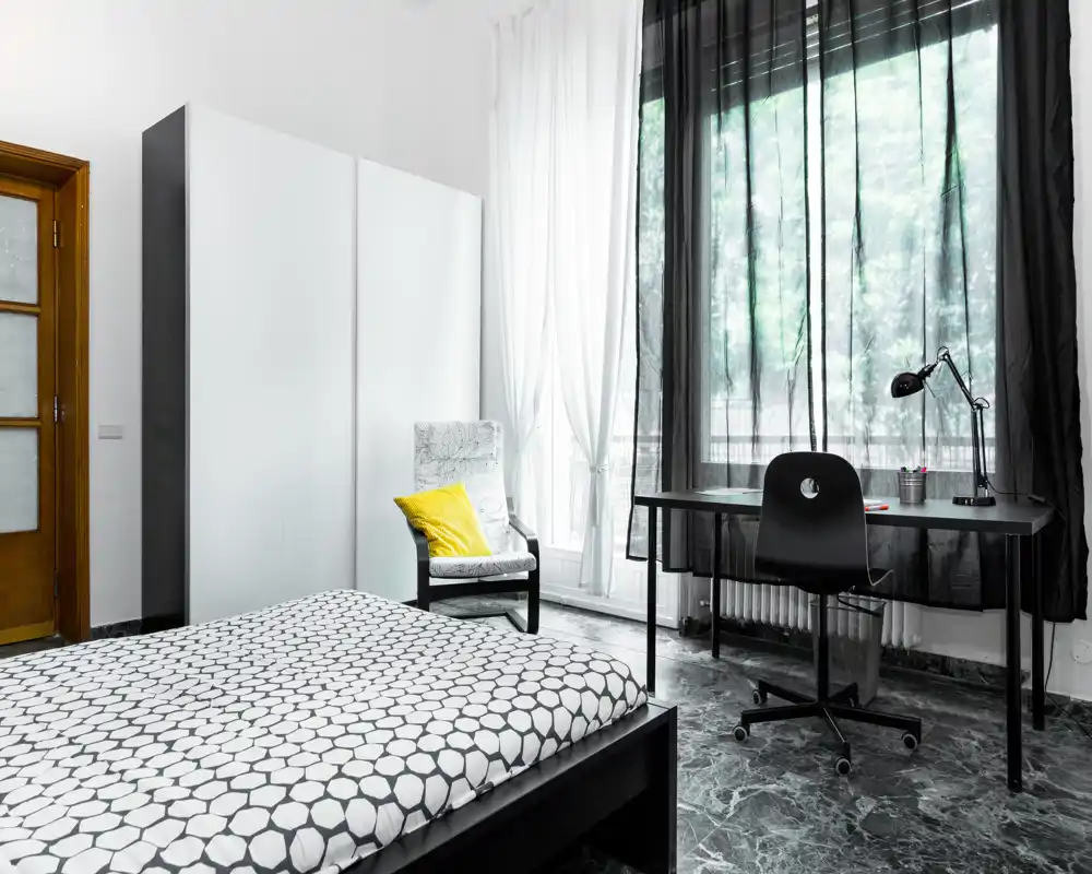 Dormitorio moderno y espacioso con cama doble estampada en primer plano, gran armario, butaca con cojín amarillo y escritorio junto a una ventana alta que aporta luz natural.