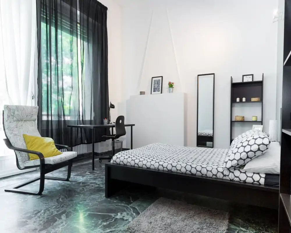 Dormitorio moderno y luminoso con cama de estampado geométrico, sillón junto a la ventana y pequeño escritorio; líneas limpias y luz natural que crean un espacio tranquilo y funcional.