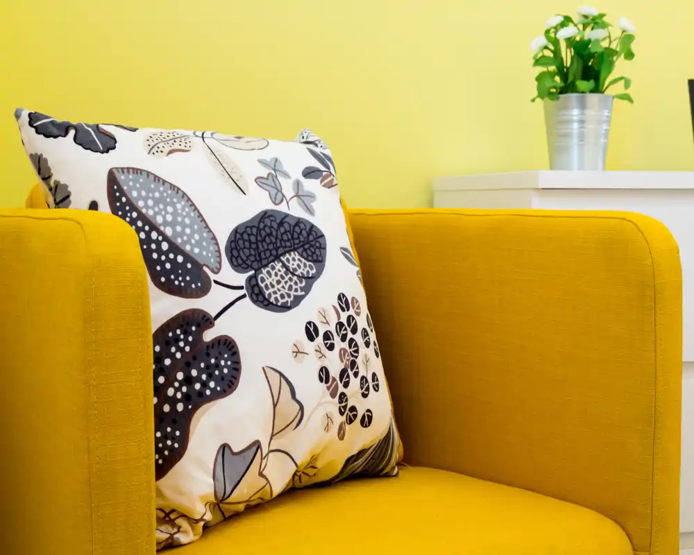 Detalle de sala luminosa y moderna: sillón color mostaza con cojín decorativo estampado y pequeña planta sobre mesita blanca — toma acogedora y con estilo perfecta para anuncios.