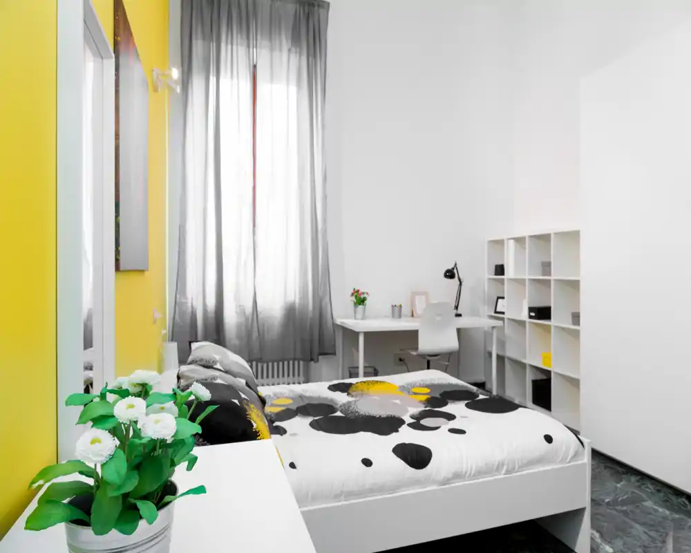 Dormitorio moderno y luminoso con cama doble estampada, escritorio y estanterías. La pared amarilla y la luz natural crean un ambiente fresco y acogedor.
