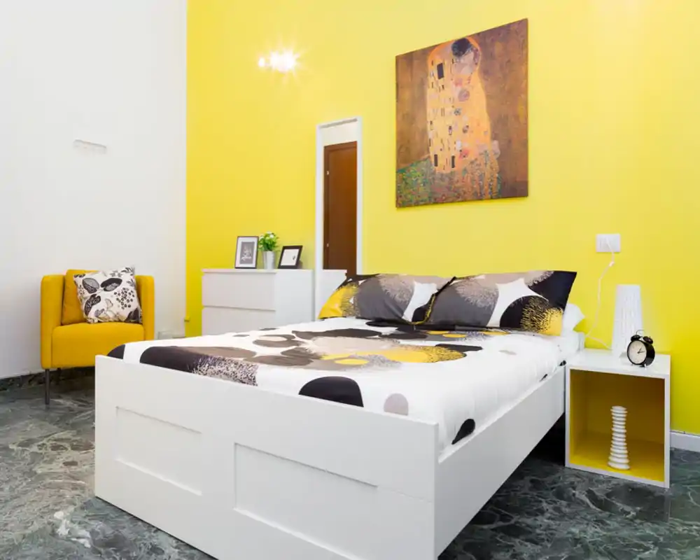 Dormitorio moderno y luminoso con cama baja blanca, ropa de cama estampada y una llamativa pared amarilla. La habitación incluye mesita, despertador, cómoda y un sillón amarillo: estilo limpio y contemporáneo, perfecto para un anuncio.
