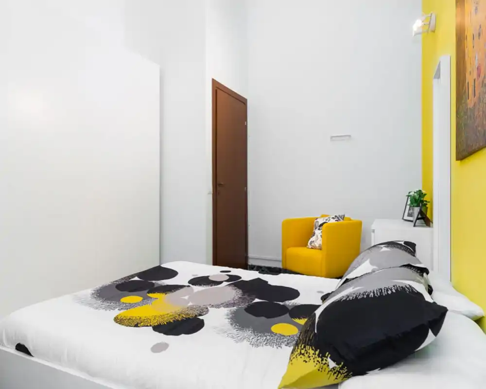 Dormitorio moderno y luminoso con cama doble estampada, cojines coordinados y un sillón amarillo como acento. Paredes blancas y mobiliario contemporáneo crean un ambiente fresco y acogedor.
