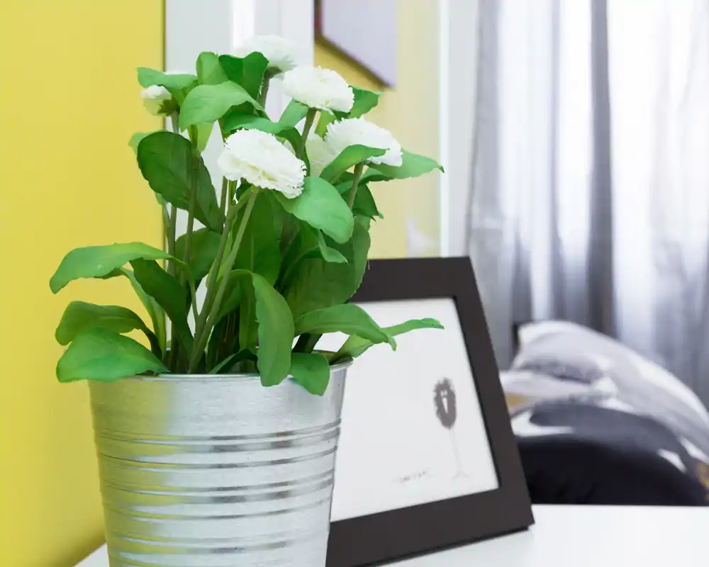 Primer plano de una mesita: una maceta metálica con flores blancas junto a un marco, con parte de la cama y las almohadas visibles al fondo.