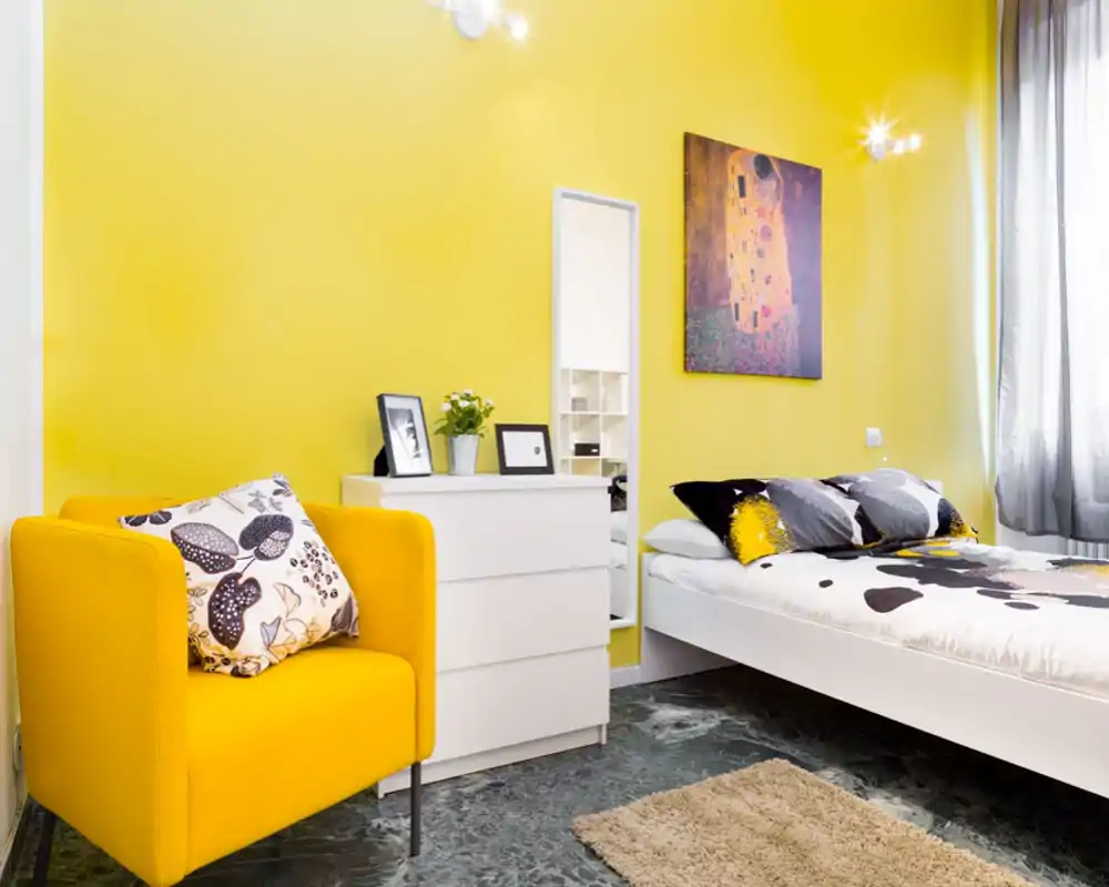 Dormitorio moderno y luminoso con cama blanca, cojines estampados y una pared amarilla intensa. Sillón amarillo a juego y cómoda blanca ofrecen un estilo fresco y atractivo para el anuncio.