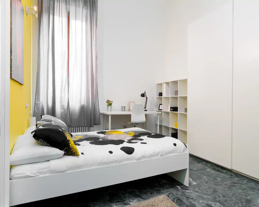 Dormitorio moderno y luminoso con cama blanca, ropa de cama estampada y un pequeño escritorio junto a la ventana. Estanterías limpias y armario amplio con un toque de color amarillo.