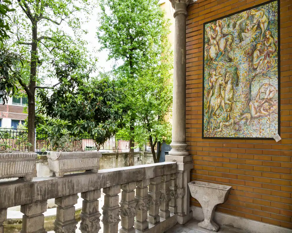 Sunny stone balcony with decorative balustrade, potted stone planters and a framed relief on a brick wall, overlooking leafy trees—ideal for a quiet outdoor corner.