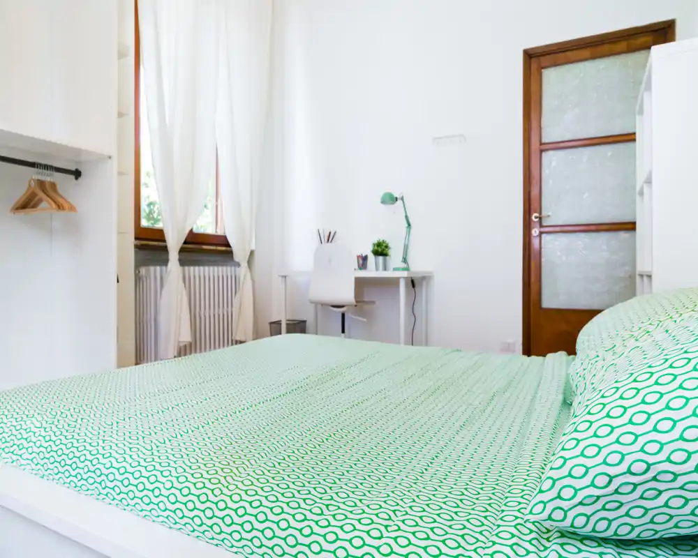 Bright, neatly staged bedroom with a green-patterned bedspread, a small white desk and chair by the window, and natural light from sheer curtains.