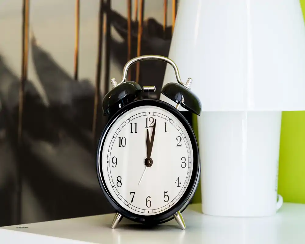 Close-up of a bedside alarm clock on a surface next to a lamp; no room layout or dominant furniture visible.