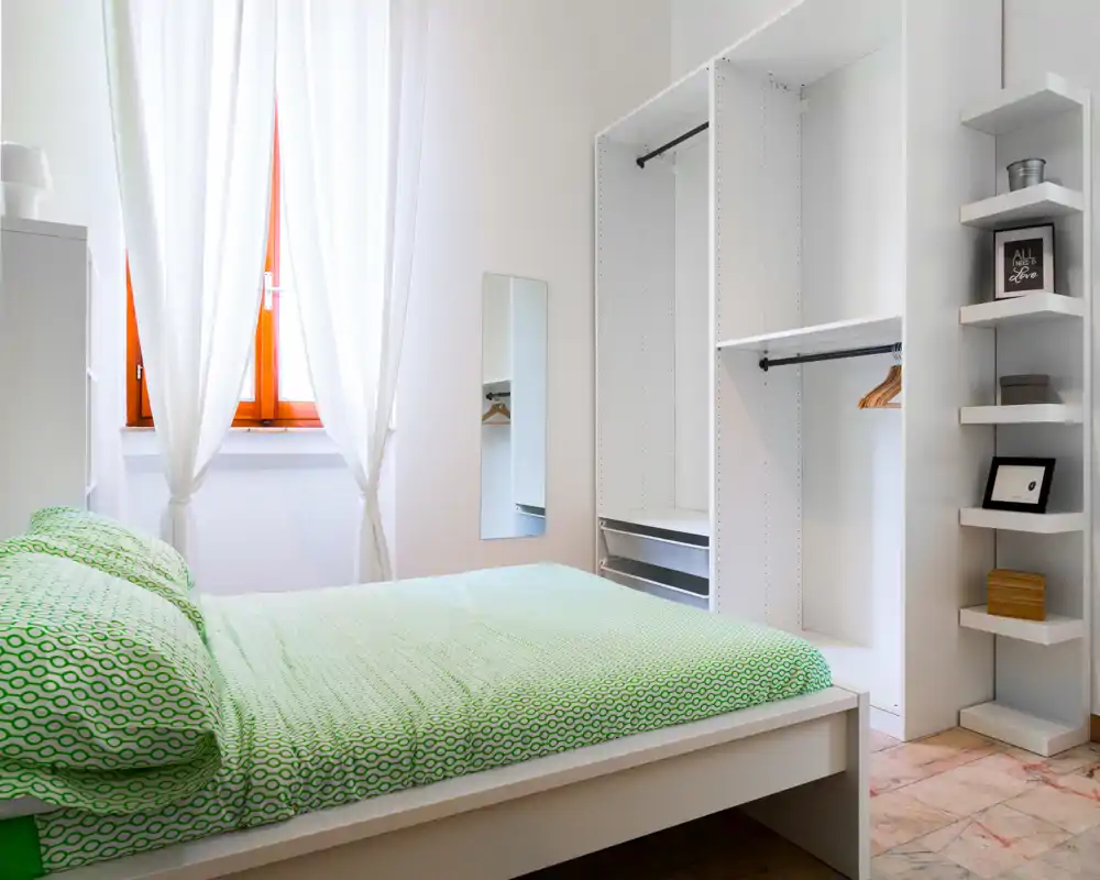 Bright, minimalist bedroom with a green-patterned bed, natural light from a window and built-in open wardrobe.