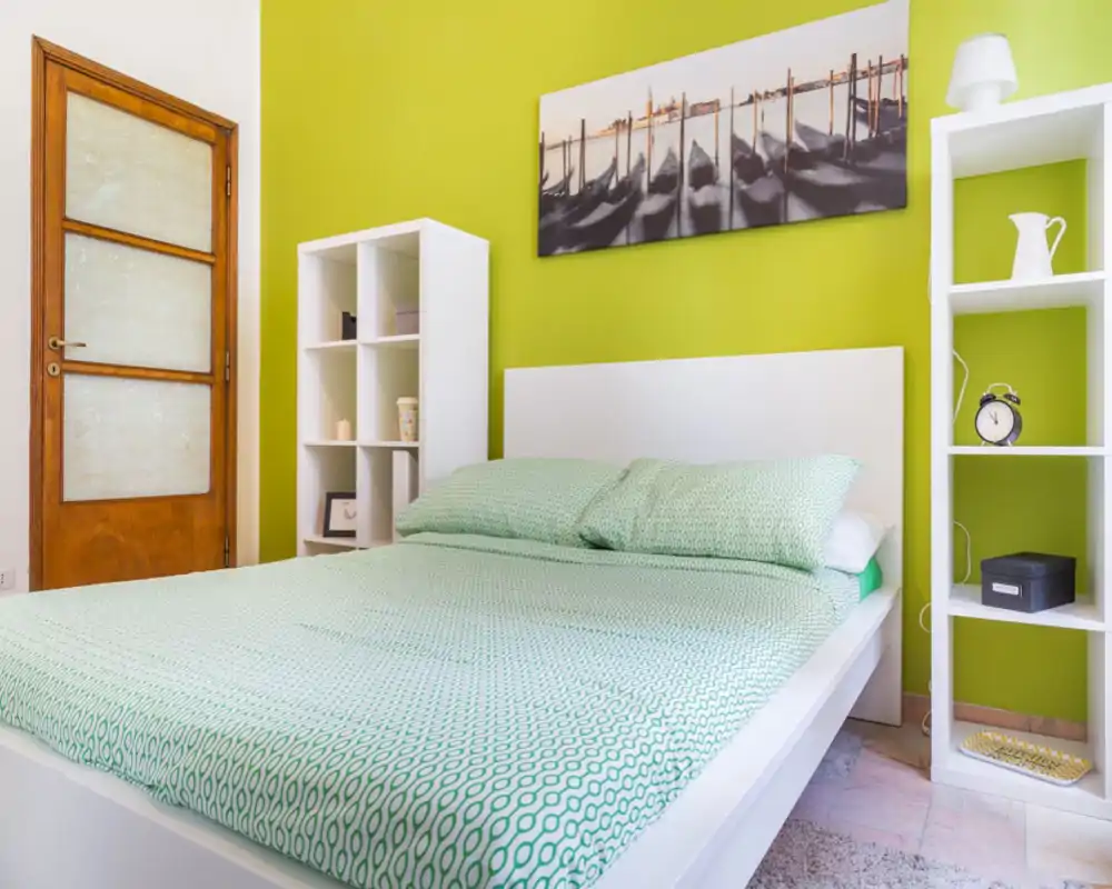 Bright, modern bedroom with a large white bed dressed in patterned mint bedding against a vivid green accent wall; simple shelving and tasteful wall art complete the cozy, well-lit space.
