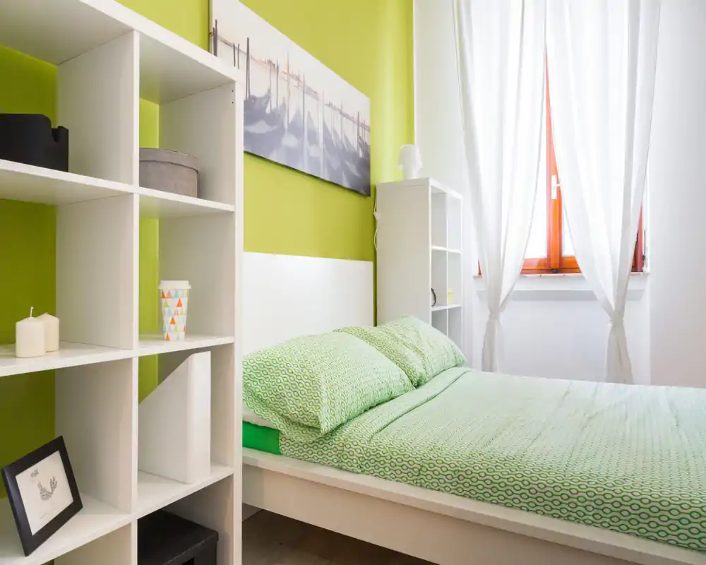 Bright, modern bedroom with green-accented décor, a neatly made double bed and white shelving beside a sunlit window.
