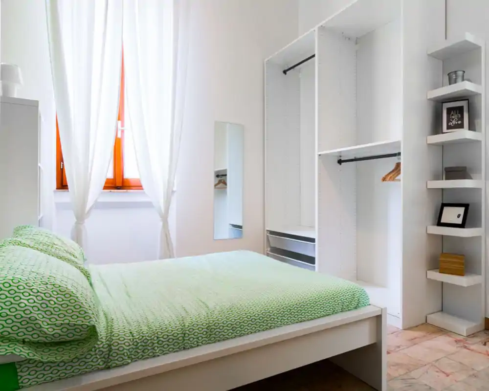 Bright, minimalist bedroom with a large white bed dressed in green patterned bedding, natural light from a curtained window, and an open wardrobe providing storage.