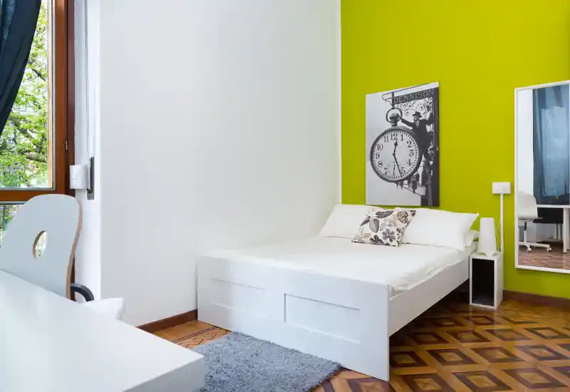 Chambre moderne et lumineuse avec un lit double blanc, un coussin décoratif et un grand miroir. La lumière naturelle et le mur vert vif donnent une ambiance fraîche et stylée, idéale pour une annonce.