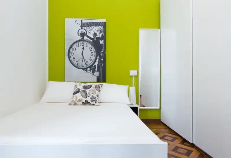 Chambre moderne et lumineuse avec lit blanc épuré, coussin décoratif et mur vert vif orné d’un tableau. Lignes nettes, parquet et armoire pleine hauteur offrent une atmosphère soignée et élégante, idéale pour une annonce locative.