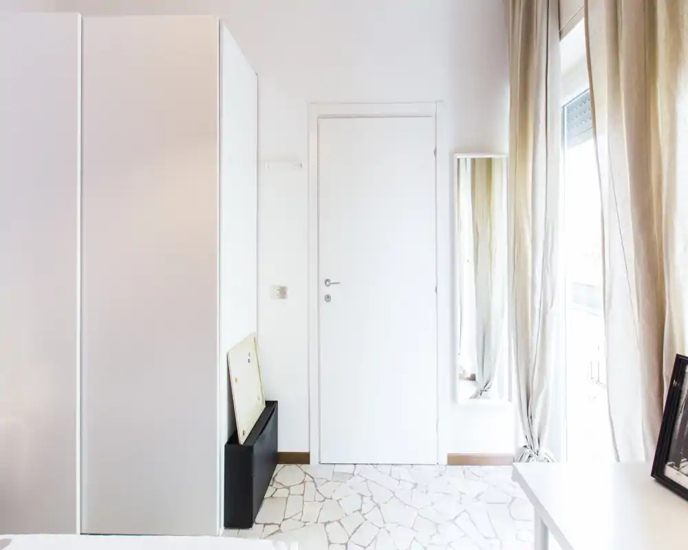 Bright, minimal room corner with wardrobe, door and window; only a small portion of the bed is visible at the lower-left. Neutral, airy styling with light curtains and terrazzo-like floor.