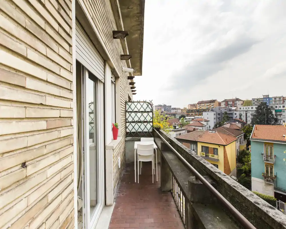 Sunny narrow balcony with a small table and two chairs, overlooking neighboring rooftops and a city view — ideal for morning coffee.