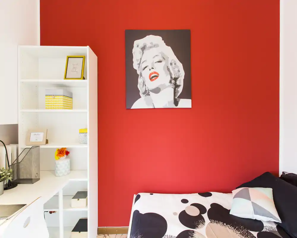 Compact bedroom with a single bed against a bold red accent wall, decorative cushions and a nearby white shelving unit offering storage and workspace.