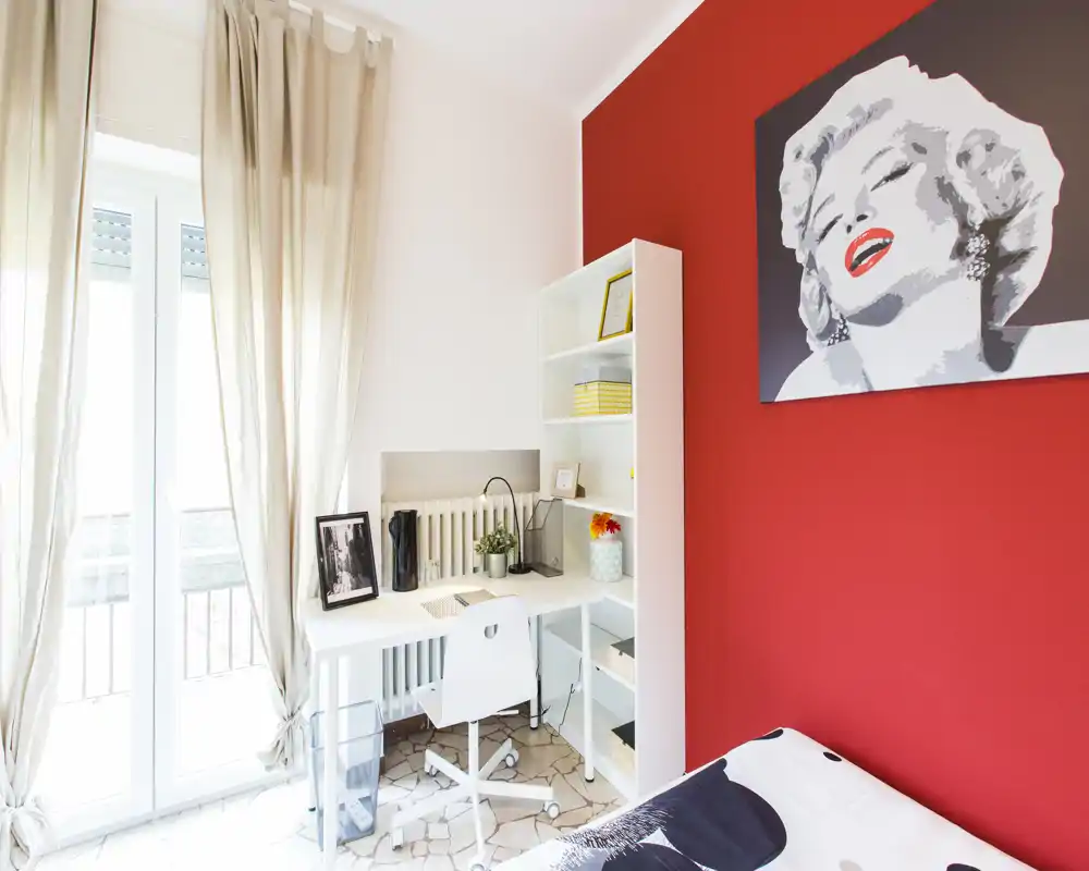 Bright, stylish bedroom corner with a white desk by a large window, cozy bedding and a bold red accent wall featuring pop-art. Natural light, neat workspace and modern decor make it ideal for listings.