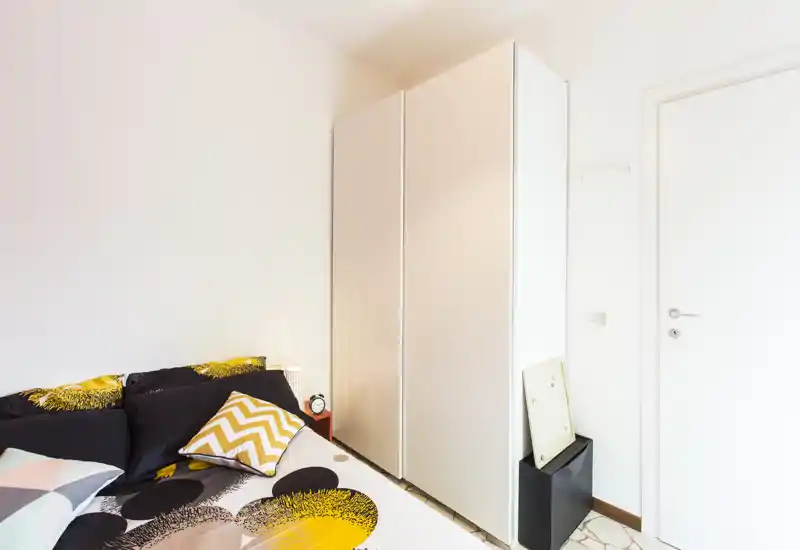 Bright, modern bedroom corner with a patterned bed, cushions and a tall white wardrobe — clean, minimal styling and pleasant natural light.