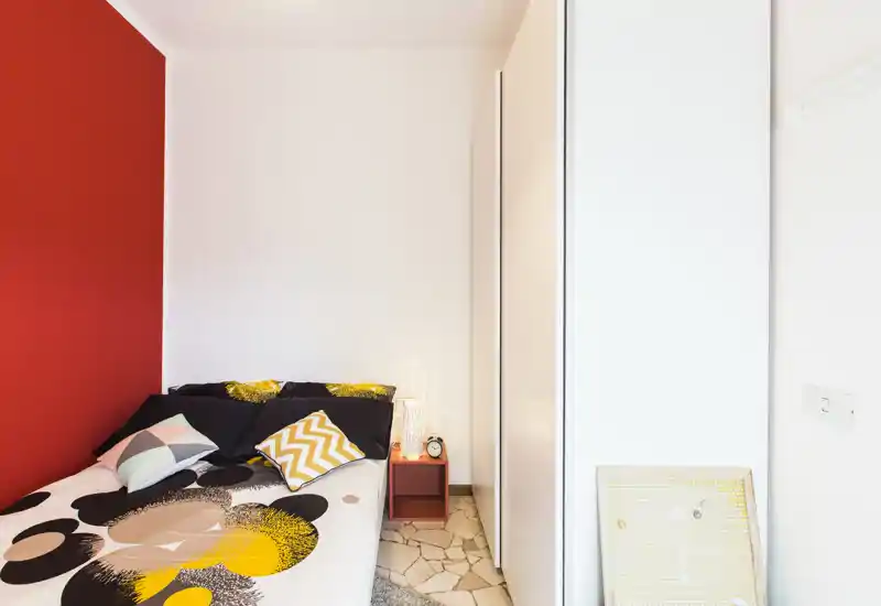 Bright, modern bedroom with a patterned duvet, colorful cushions and a red accent wall. The room features a bedside table with a lamp and a compact wardrobe—ideal for a stylish single or couple.