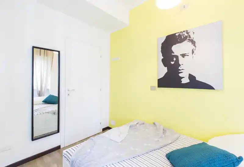 Camera da letto luminosa e moderna con letto a righe, cuscino turchese e un grande ritratto sulla parete gialla. La stanza appare ariosa grazie alla luce naturale e a un arredo essenziale e contemporaneo.