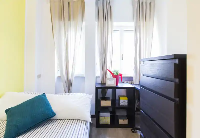 Camera da letto luminosa e moderna con letto ben rifatto, cuscino turchese e un comò in legno scuro accanto a una nicchia con finestra.