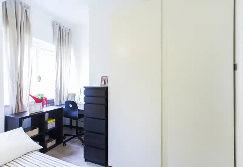 Camera da letto luminosa e minimale con angolo del letto visibile, armadio con ante scorrevoli e una scrivania compatta sotto la finestra — stile moderno e pulito, ideale per una persona.