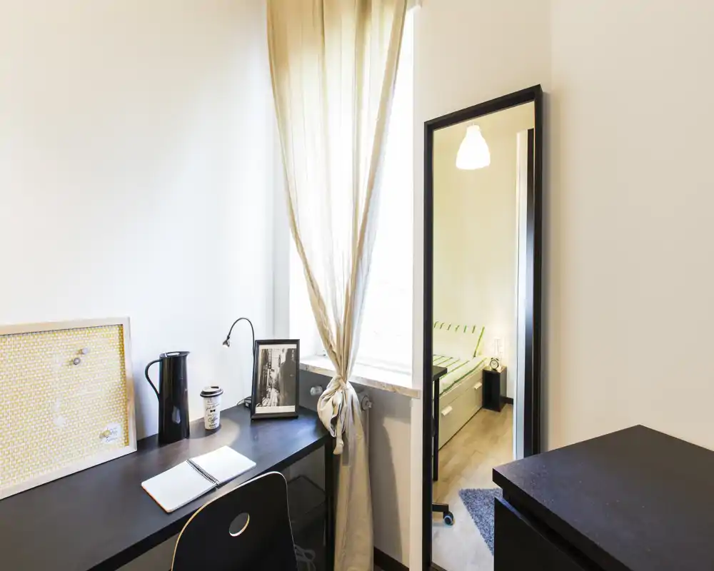 Compact bedroom corner shown through a doorway mirror: a tidy desk by a bright window and the bed visible as a reflection. Natural light, simple modern decor and functional workspace make it suitable for a single occupant.