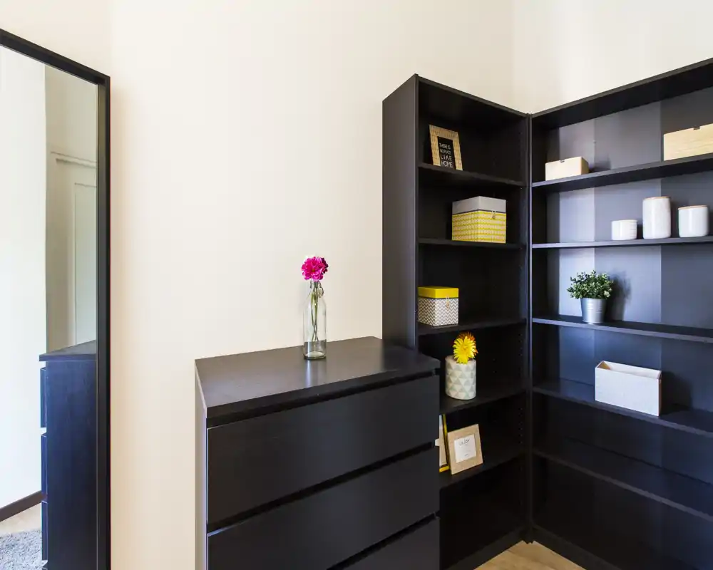 A tidy bedroom corner with dark wooden dresser and matching shelving, decorative boxes and a vase with fresh flowers; bright, minimal and well organized.