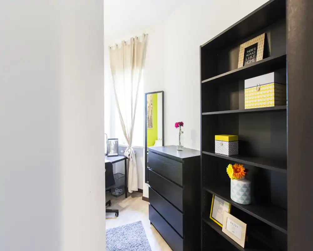 Small, bright bedroom corner with a black dresser, open shelving and a desk by the window; neutral curtains and decorative accents create a neat, functional space.