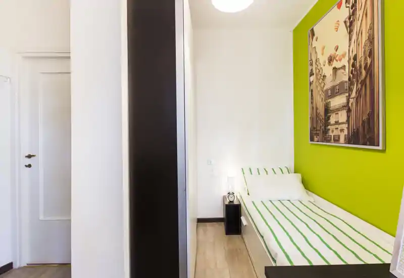 Compact single bedroom with a neatly made bed against a bright green accent wall, bedside lamp and framed city artwork; clean lines and warm wooden flooring create a fresh, welcoming space.