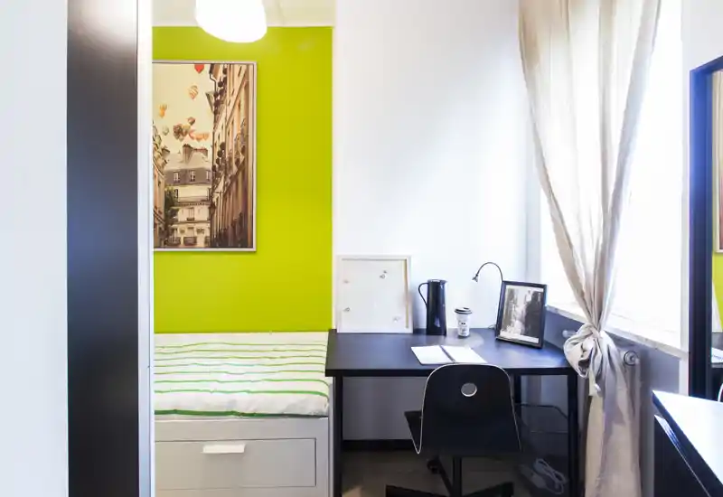 Compact staged bedroom with a single bed against a vibrant green accent wall and a tidy workspace by a bright window — ideal for a student or single occupant.