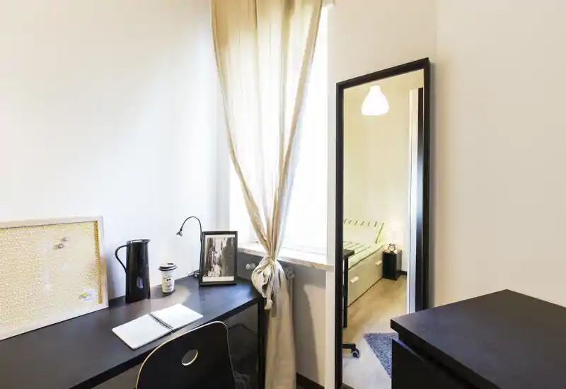 Compact bedroom corner shown through a doorway mirror: a tidy desk by a bright window and the bed visible as a reflection. Natural light, simple modern decor and functional workspace make it suitable for a single occupant.