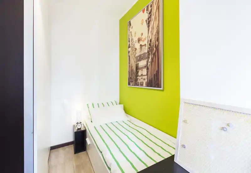 Compact single bedroom with a neat single bed against a vibrant green accent wall, framed artwork and a small bedside table — bright and well styled for a cozy stay.