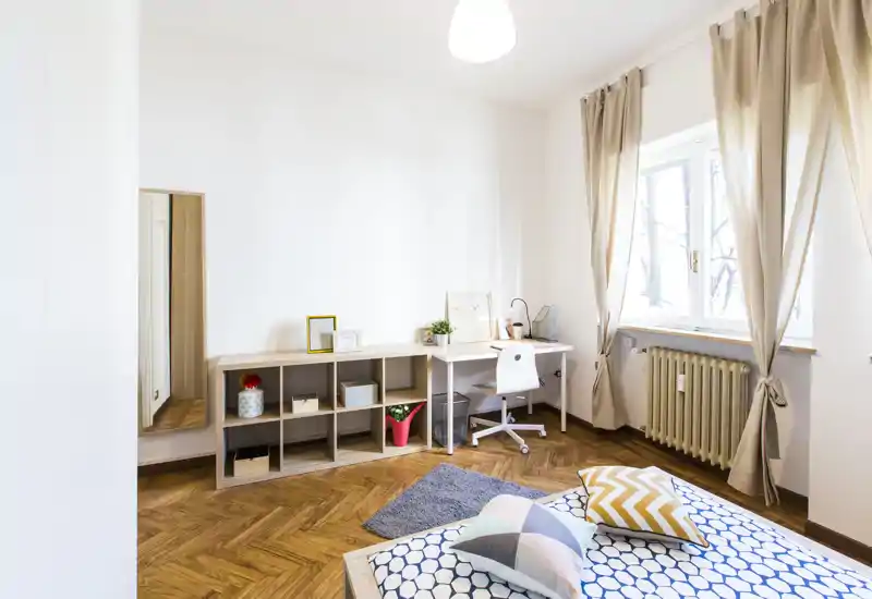 Bright, modern bedroom with wooden parquet, patterned bedding and a small workspace by the window — airy and well styled for rental listings.