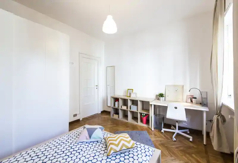 Bright, modern bedroom with a patterned bed, decorative cushions and a tidy desk by the window — clean parquet flooring and minimalist storage create a calm, welcoming space.