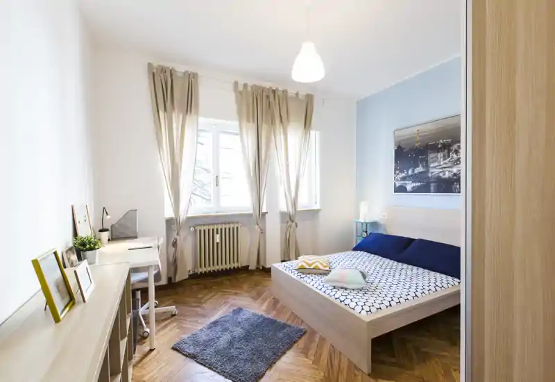 Bright, modern bedroom with a double bed, decorative cushions and a small desk by the window. Hardwood floors, soft rug and neutral curtains create a welcoming, well-lit space.