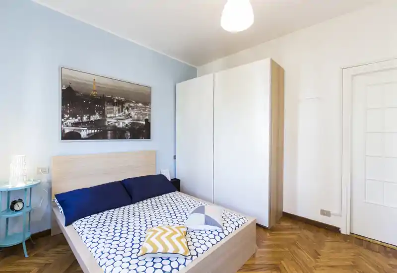Bright, stylish bedroom with a double bed, patterned bedding and cozy decorative cushions; white wardrobe and soft blue accent wall create a calm, modern atmosphere.