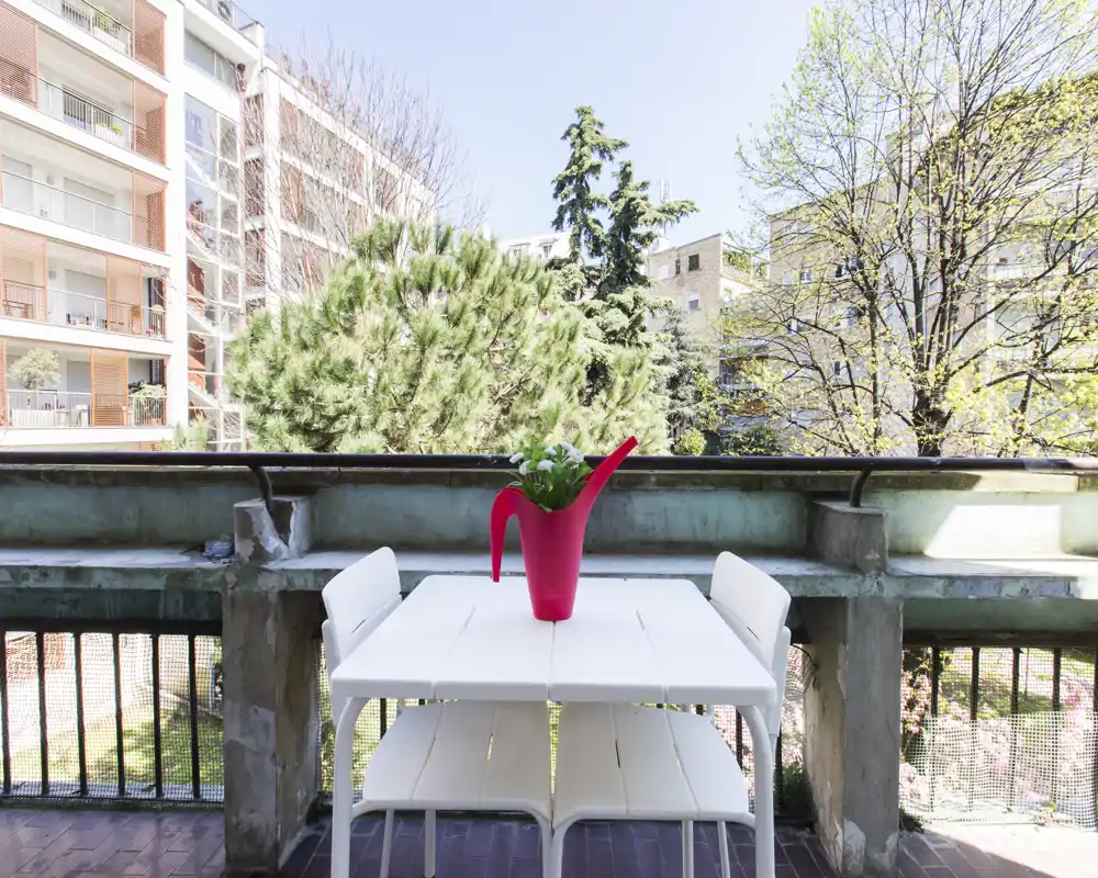 Sunny balcony with a white table and chairs and a red watering-can planter, overlooking trees and neighboring buildings — ideal for morning coffee.