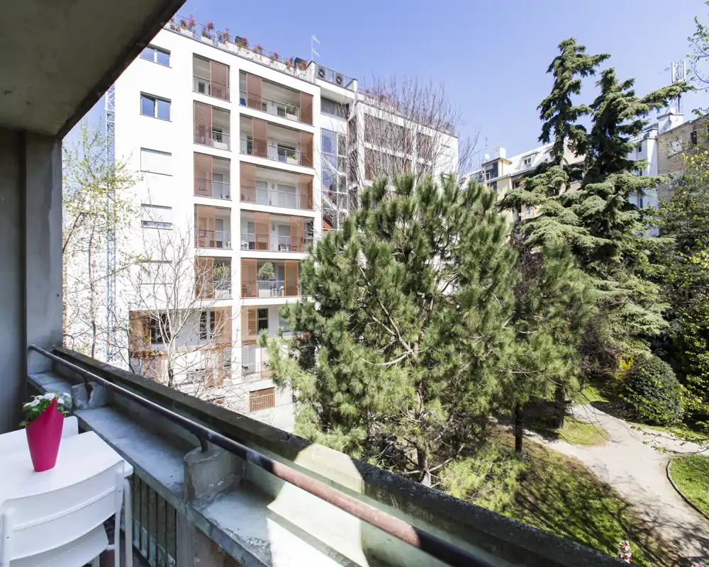 Sunny private balcony overlooking a leafy courtyard and neighboring modern building — small table and chairs perfect for morning coffee.
