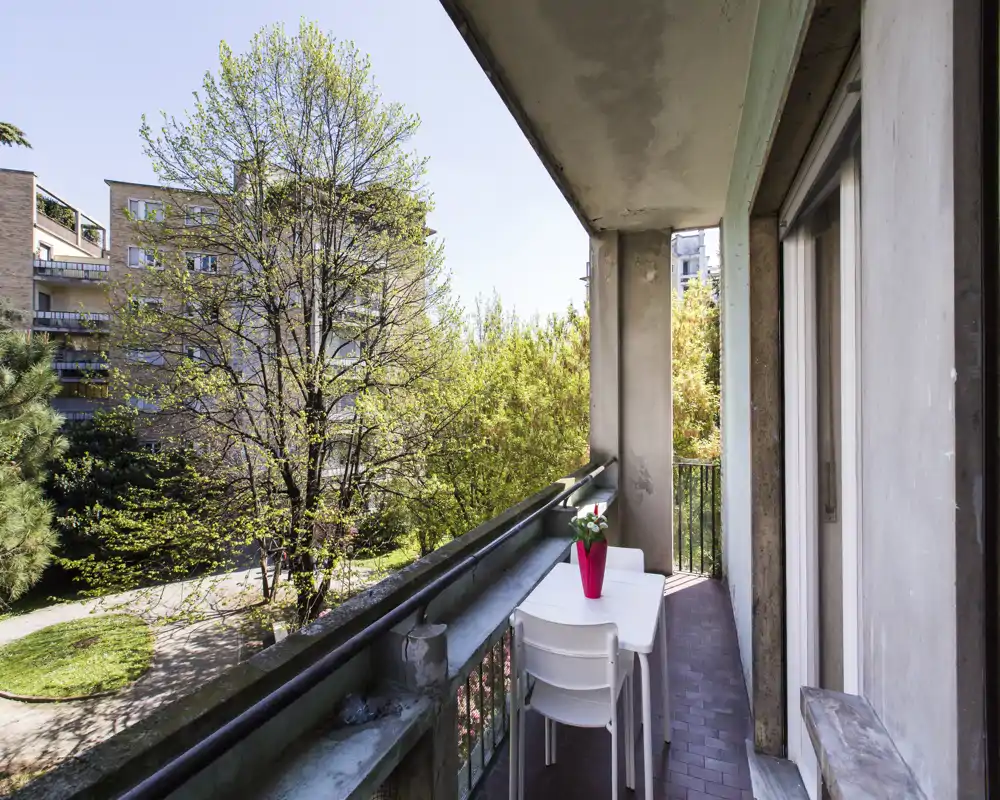 Sunny balcony with a small white table and two chairs, overlooking leafy trees and neighboring buildings — a pleasant outdoor spot for morning coffee.
