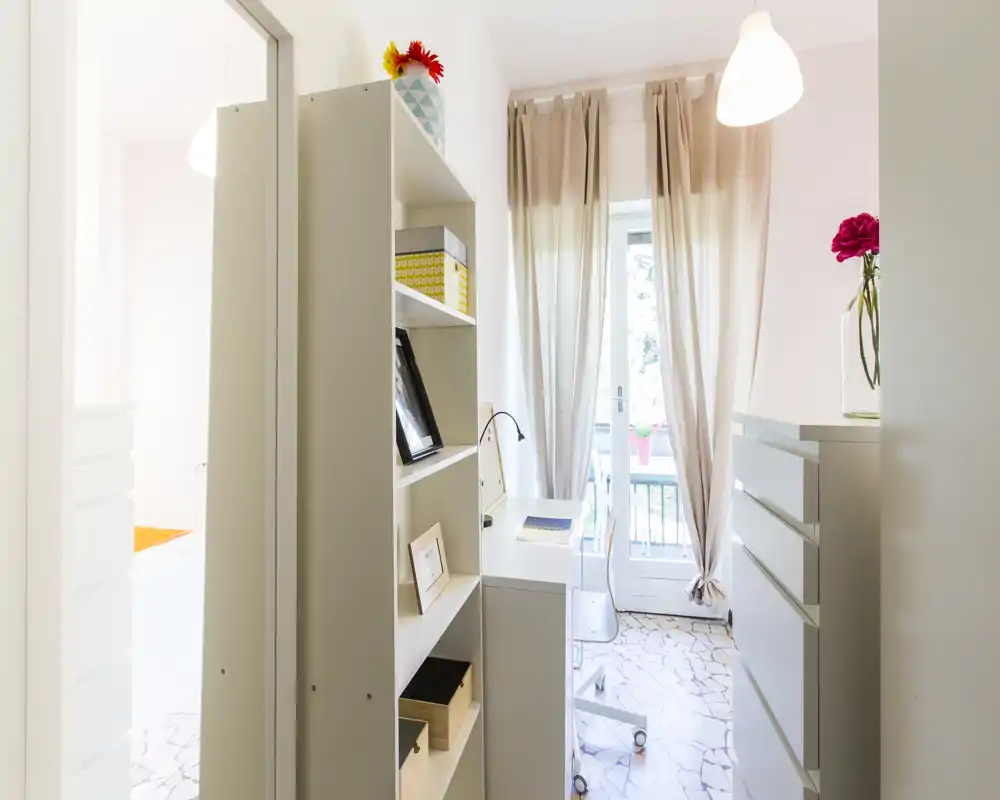 Bright, neatly styled living/working corner with a shelving unit, desk and balcony door; soft curtains and decorative touches create a welcoming atmosphere.