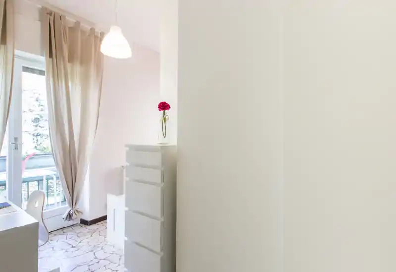 Bright, small bedroom corner with a chest of drawers, desk and balcony access; light curtains and a hanging lamp give an airy feel.