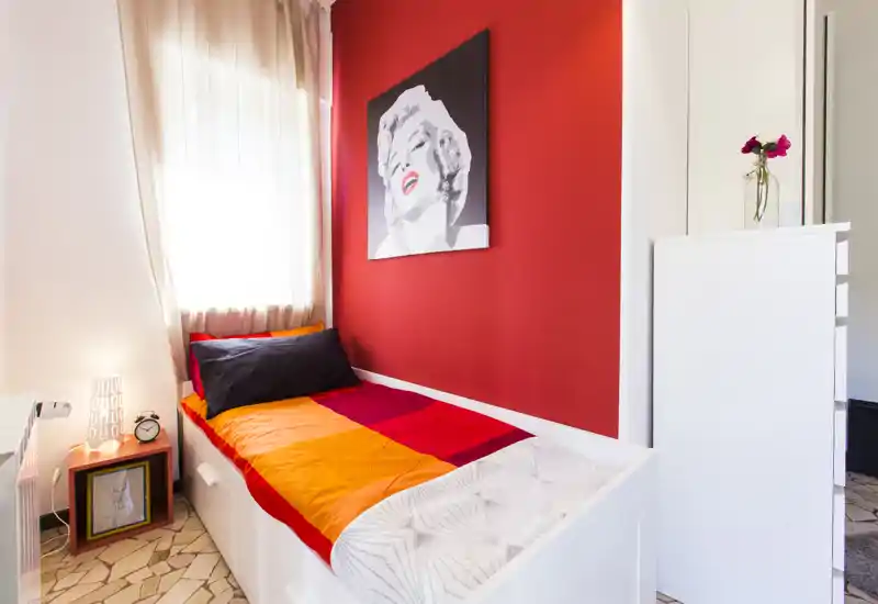 Compact single bedroom with a bold red accent wall, a neatly made single bed with colorful bedding, a bedside lamp and framed artwork — bright and well-styled for a cozy stay.
