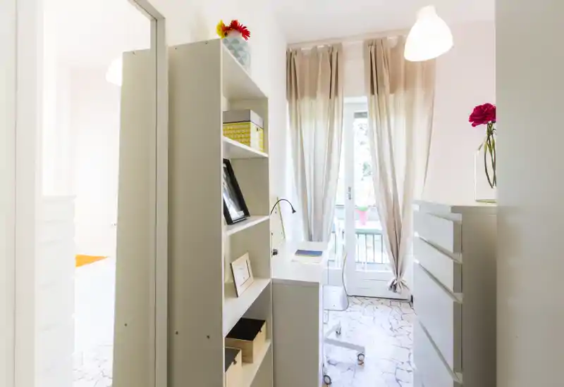 Bright, neatly styled living/working corner with a shelving unit, desk and balcony door; soft curtains and decorative touches create a welcoming atmosphere.