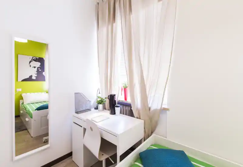 Camera luminosa e compatta con scrivania bianca vicino alla finestra, biancheria a righe verdi e cuscino blu. Ben illuminata da luce naturale e arredata in modo ordinato per un soggiorno singolo confortevole.