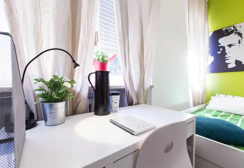 Angolo camera luminoso e moderno con una scrivania bianca in primo piano, pianta decorativa e lampada, e un letto ordinato con biancheria verde su una parete accentata color lime.