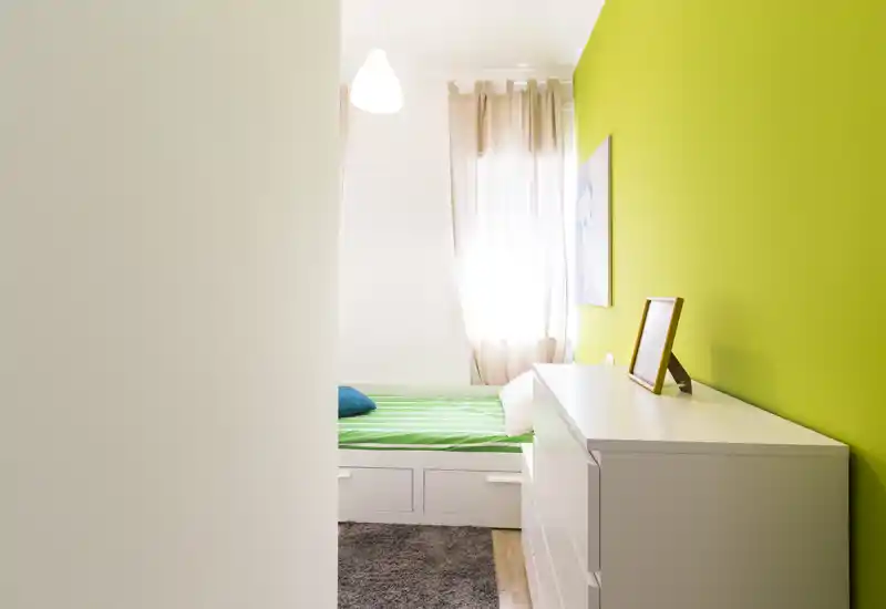Camera singola compatta e luminosa con parete verde, letto con cassetti integrati e comò bianco; la luce naturale dalla finestra crea un'atmosfera fresca e accogliente.