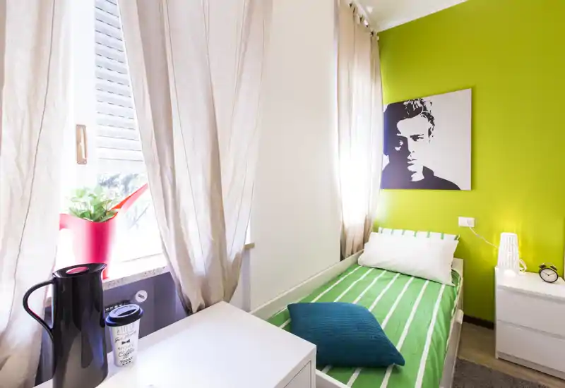 Camera da letto piccola e luminosa con singolo letto con copriletto verde a righe, cuscino turchese e comodino con lampada. La luce naturale dalla finestra e l'opera murale moderna rendono l'ambiente fresco e accogliente.